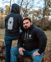 A man outdoors wears a black Farm Fresh Clothing hoodie with a white cow logo, ideal for food freedom and raw milk fans. Next to him, another person sports the Raw is Law Hoodie featuring a jar graphic and the phrase “How Is Lunch?”.