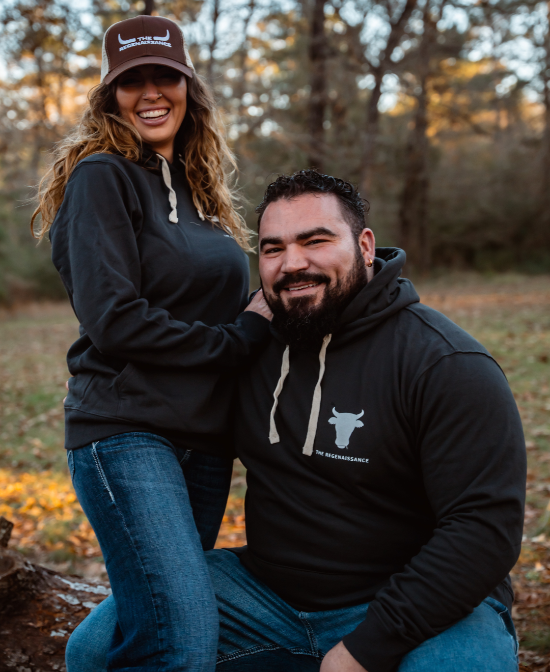 A man and woman outdoors amid autumn trees wear black Farm Fresh Clothing Raw is Law Hoodies with white bull logos. Smiling, she stands with a hand on his shoulder as he sits on a log, both enjoying the scenic backdrop.