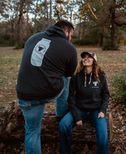 A man and woman outdoors in jeans wear Farm Fresh Clothing’s Raw is Law Hoodie. The man stands with a knee on a log, showing off the bold logo, while the woman sits beside him, smiling.