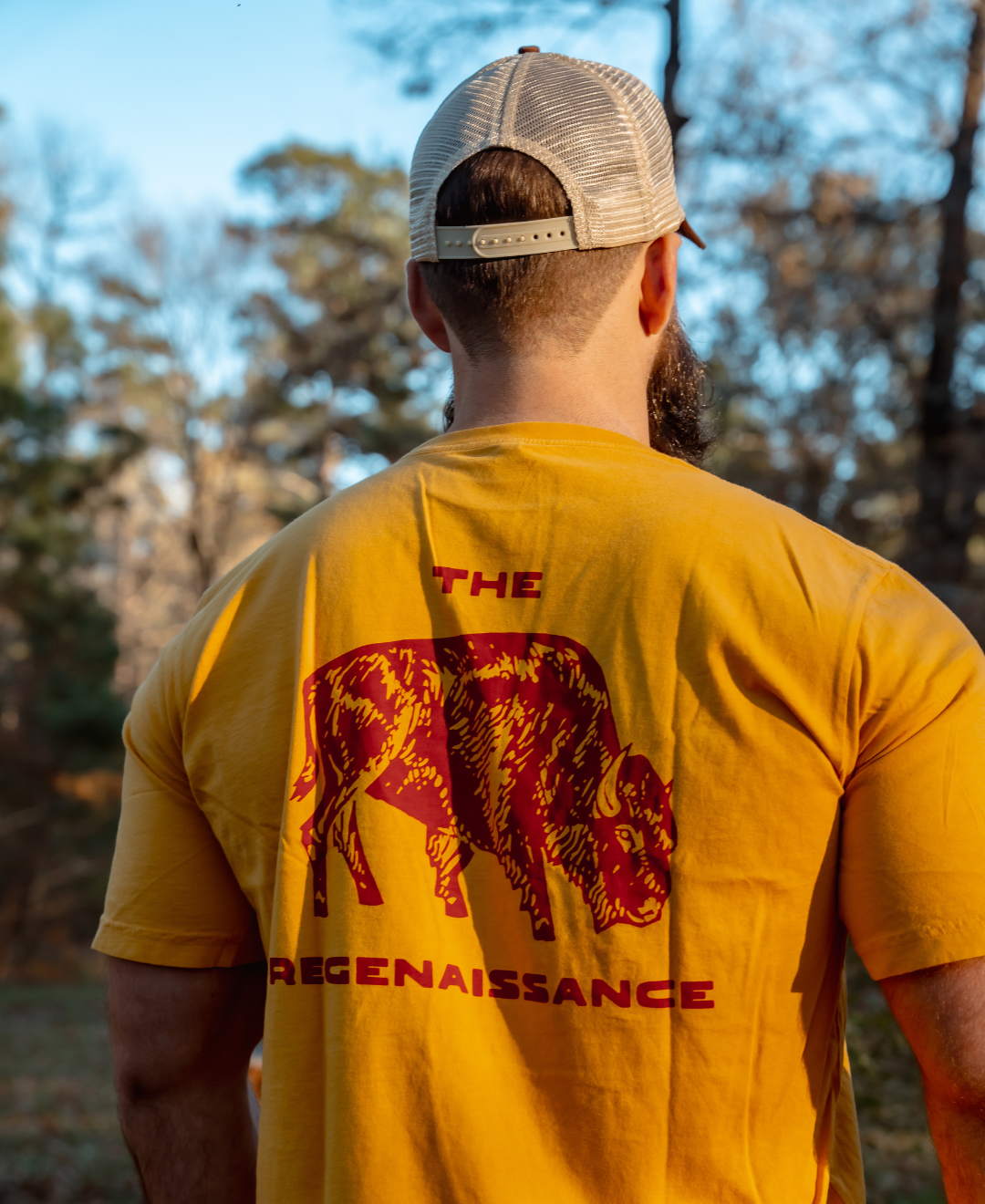A man stands outdoors, facing away in sunlight and trees, wearing a yellow shirt with a red bison graphic and “THE REGENAISSANCE,” paired with Farm Fresh Clothings Longhorn Trucker Hat celebrating food freedom.