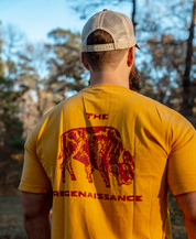 A man stands outdoors, facing away in sunlight and trees, wearing a yellow shirt with a red bison graphic and “THE REGENAISSANCE,” paired with Farm Fresh Clothings Longhorn Trucker Hat celebrating food freedom.