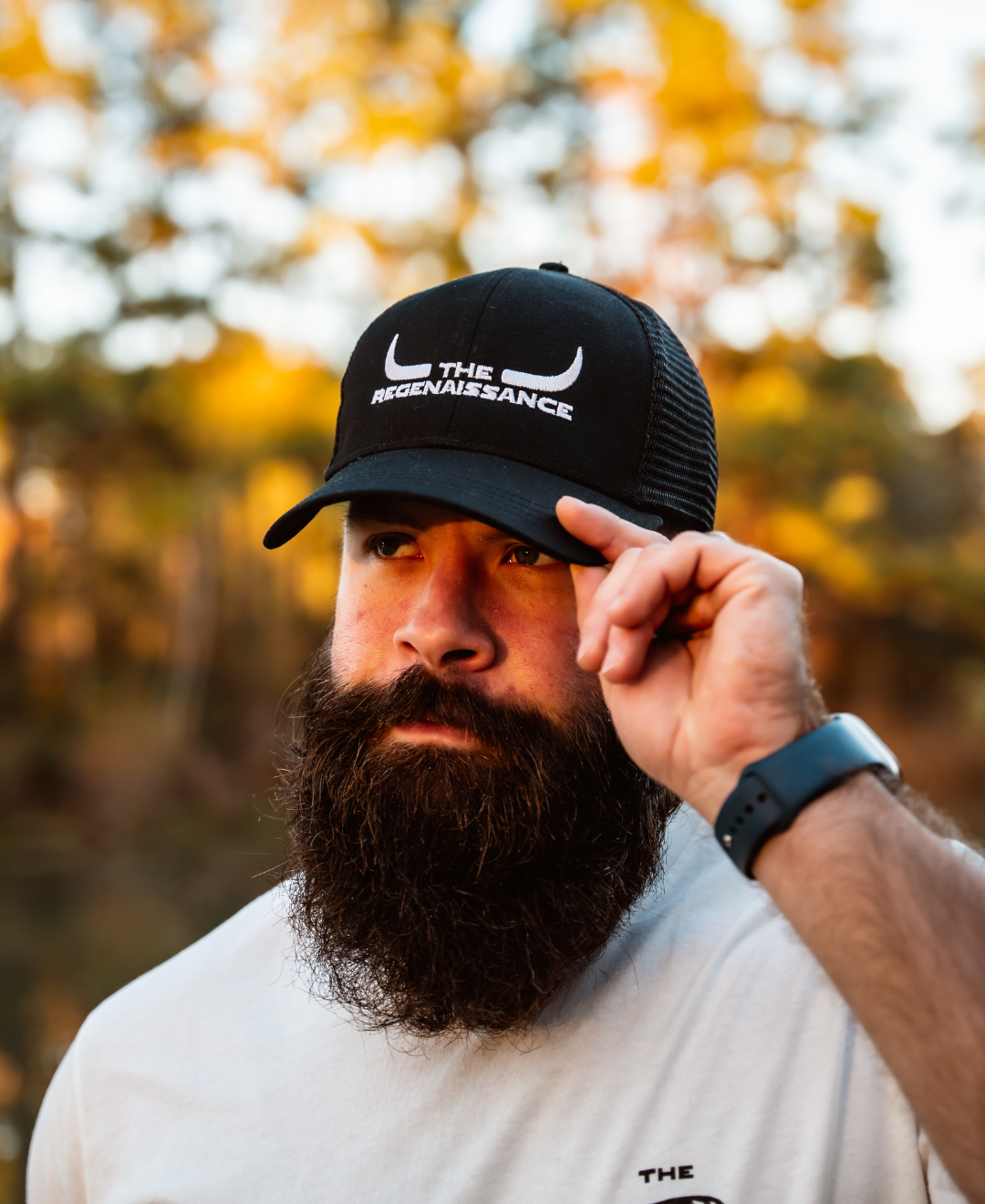 A bearded man in a white shirt and a black/black Longhorn Trucker Hat from Farm Fresh Clothing stands outdoors, holding the brim of his hat. Trees and sunlight in the blurry background evoke the spirit of food freedom.