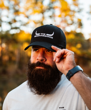 A bearded man in a white shirt and a black/black Longhorn Trucker Hat from Farm Fresh Clothing stands outdoors, holding the brim of his hat. Trees and sunlight in the blurry background evoke the spirit of food freedom.