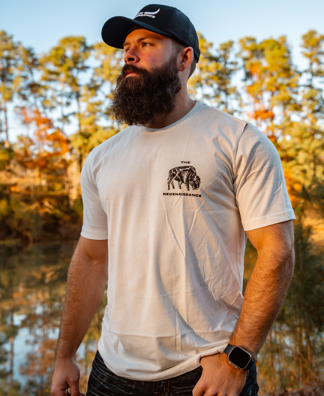 A bearded man in a Farm Fresh Clothing Longhorn Trucker Hat, white T-shirt with The Recerenaissance elephant graphic, and smartwatch stands outdoors by trees and water in warm sunlight.
