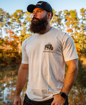 A bearded man in a Farm Fresh Clothing Longhorn Trucker Hat, white T-shirt with The Recerenaissance elephant graphic, and smartwatch stands outdoors by trees and water in warm sunlight.