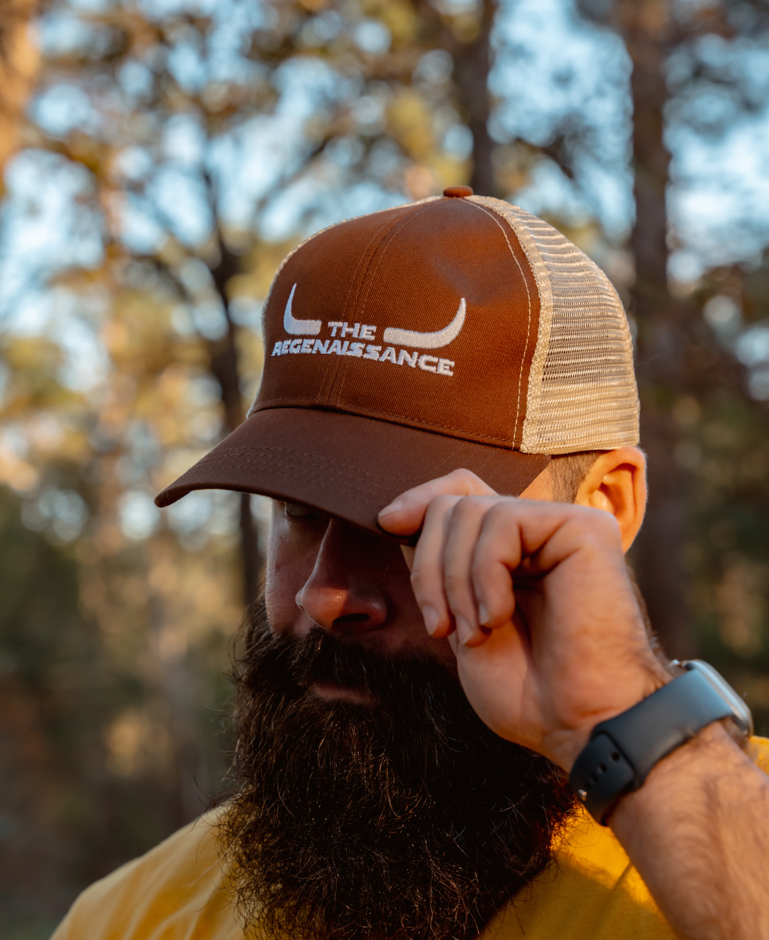A bearded person in a yellow shirt and smartwatch adjusts a Farm Fresh Clothing Longhorn Trucker Hat in Earth/Oyster, featuring white bull horns and THE RENAISSANCE text, standing outdoors with blurred trees in the background.