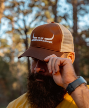 A bearded person in a yellow shirt and smartwatch adjusts a Farm Fresh Clothing Longhorn Trucker Hat in Earth/Oyster, featuring white bull horns and THE RENAISSANCE text, standing outdoors with blurred trees in the background.