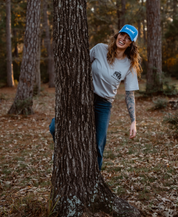 A smiling woman in a blue Farm Fresh Clothing Longhorn Trucker Hat, gray tee, and jeans playfully leans out from behind a tree amid fallen leaves, embracing food freedom.