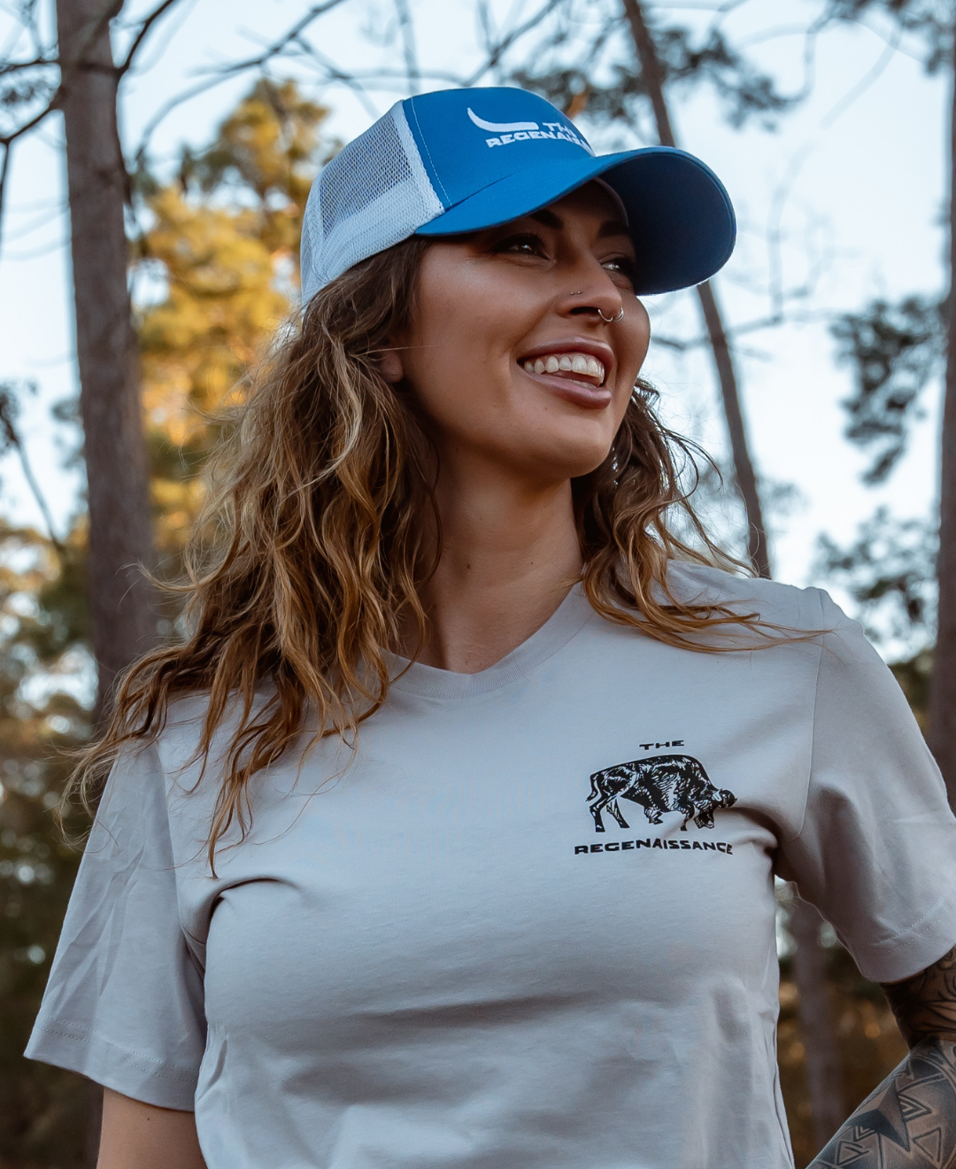A smiling woman with wavy hair wears a Farm Fresh Clothing Longhorn Trucker Hat in Daylight Blue/White and a gray T-shirt with “The Regenaissance” bull graphic, celebrating food freedom outdoors among trees and sunlight.