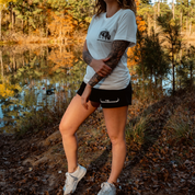 A woman with a tattoo sleeve stands by a forest pond in autumn, wearing black Los Angeles Apparel Womens Longhorn Shorts, a white t-shirt, and white sneakers. Colorful trees and leaves fill the background.