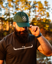 A bearded man in a spruce MMRA Trinity Cap by Farm Fresh Clothing and a The Regenaissance t-shirt stands outdoors, tilting his cap downward amid trees and warm sunlight, capturing the essence of food freedom.