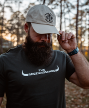 A bearded man outdoors wears a dark shirt and a light stone MMRA Trinity Cap by Farm Fresh Clothing, holding the cap’s brim with one hand. Blurred trees are visible in the background.