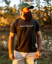 A bearded man in a dark THE REGENAISSANCE tee, white shorts, and a Farm Fresh Clothing MMRA Trinity Cap stands outdoors by a pond amid trees and soft golden sunlight.