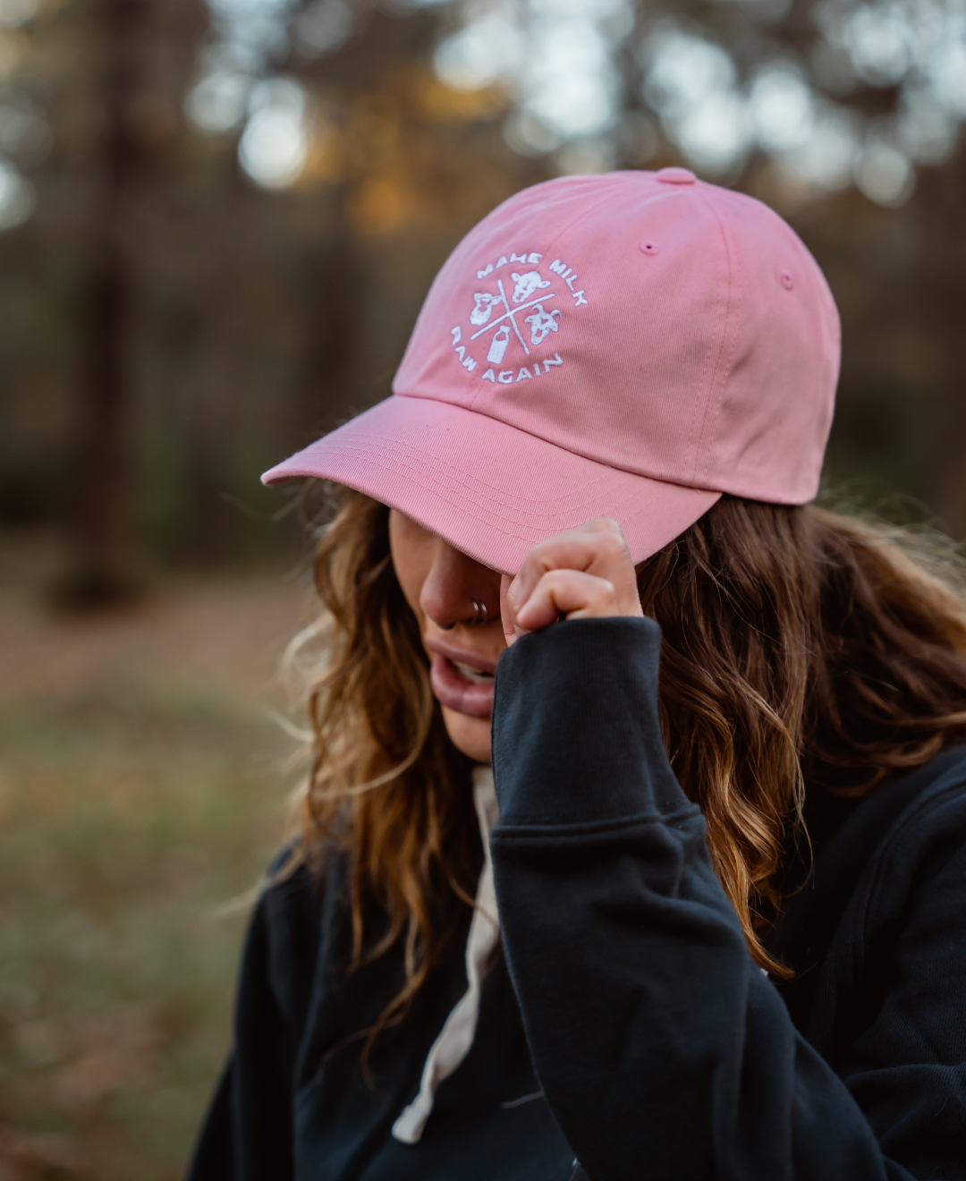 A wavy-haired person with a nose ring wears a pink Farm Fresh Clothing MMRA Trinity Cap and a dark hoodie, tilting the cap to partially hide their face while standing outdoors in a blurred wooded area.