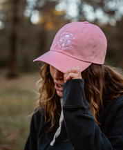 A wavy-haired person with a nose ring wears a pink Farm Fresh Clothing MMRA Trinity Cap and a dark hoodie, tilting the cap to partially hide their face while standing outdoors in a blurred wooded area.