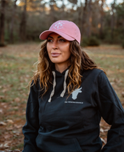 A woman with wavy brown hair sits outdoors in a Farm Fresh Clothing MMRA Trinity Cap and a black hoodie with THE REGENAISSANCE text. Blurred trees and grass in the background evoke food freedom.
