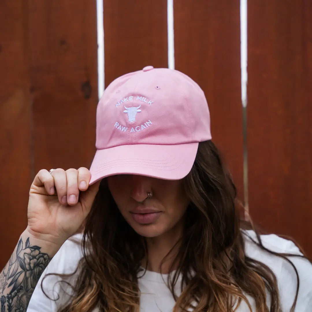 A woman with long brown hair and a nose ring wears a pink Farm Fresh Clothing MMRA Cow Cap reading "MAKE MILK RAW AGAIN," holding the brim to show her left forearm tattoo, with a wooden fence in the background, supporting food freedom.