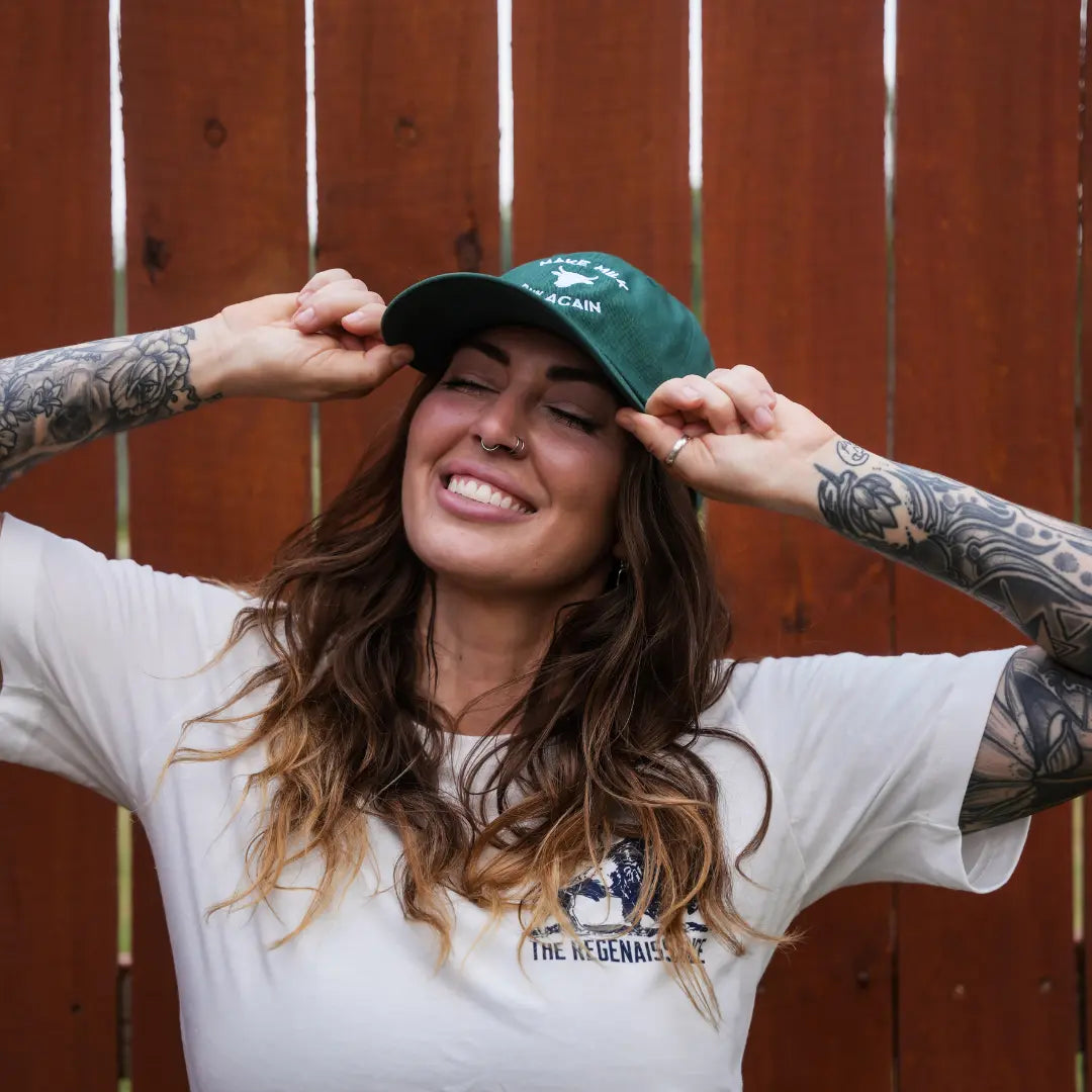 A smiling woman with long wavy hair and tattooed arms adjusts her Farm Fresh Clothing MMRA Cow Cap while standing by a wooden fence, dressed in a white t-shirt.