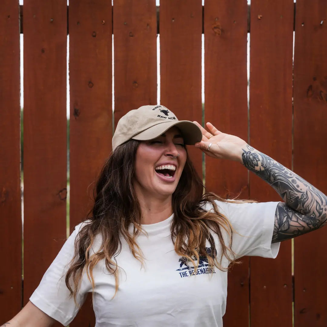 A woman with long hair and a tattooed arm laughs while holding her beige MMRA Cow Cap by Farm Fresh Clothing, wearing a white t-shirt and standing in front of a wooden fence, capturing the spirit of food freedom.