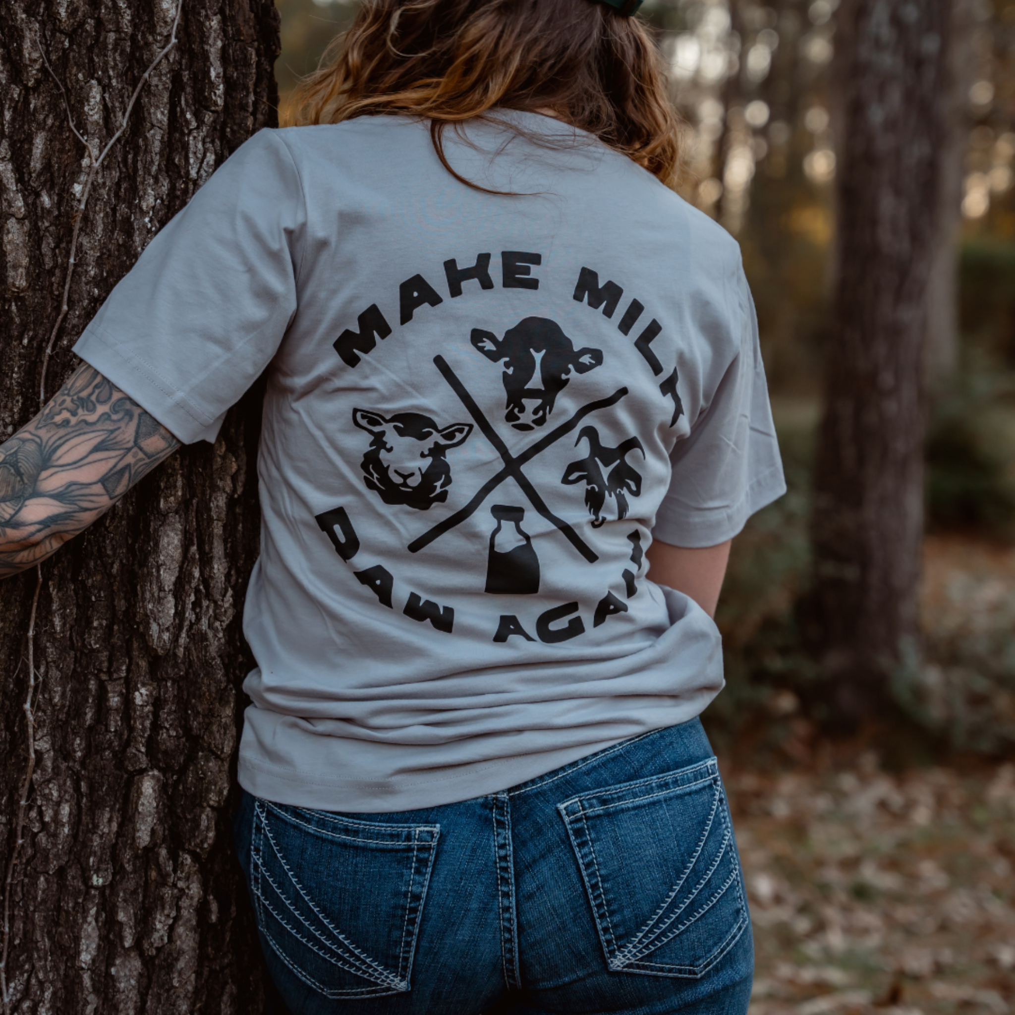 A tattooed, long-haired person leans against a tree in the forest wearing Farm Fresh Clothings MMRA Trinity Tee—an organic cotton gray shirt featuring cartoon animals, a milk bottle, and MAKE MILK RAW AGAIN on the back.