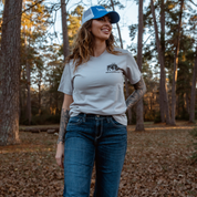 A tattooed woman in a blue cap, jeans, and the Farm Fresh Clothing MMRA Trinity Tee in Ice smiles outdoors amid fallen leaves in a forested area.