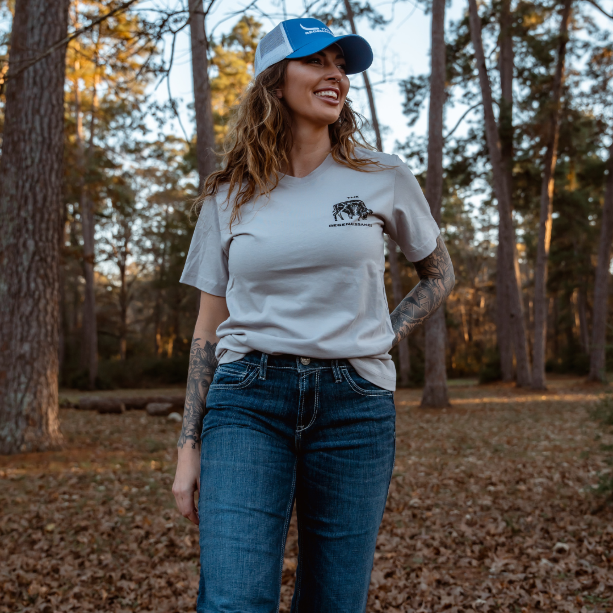 A tattooed woman in a blue cap, jeans, and the Farm Fresh Clothing MMRA Trinity Tee in Ice smiles outdoors amid fallen leaves in a forested area.