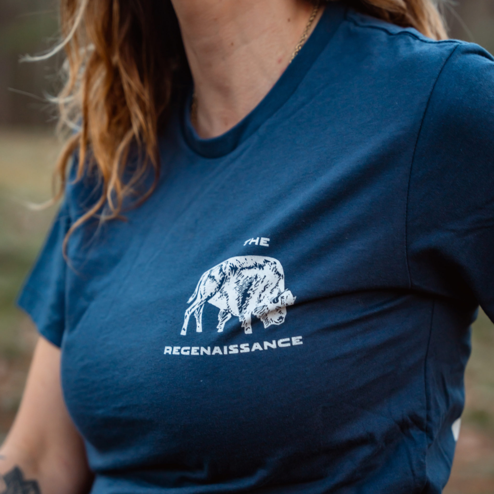 Someone with wavy hair wears the Farm Fresh Clothing MMRA Trinity Tee, a blue organic cotton t-shirt with a white bison graphic and THE REGENAISSANCE printed below.