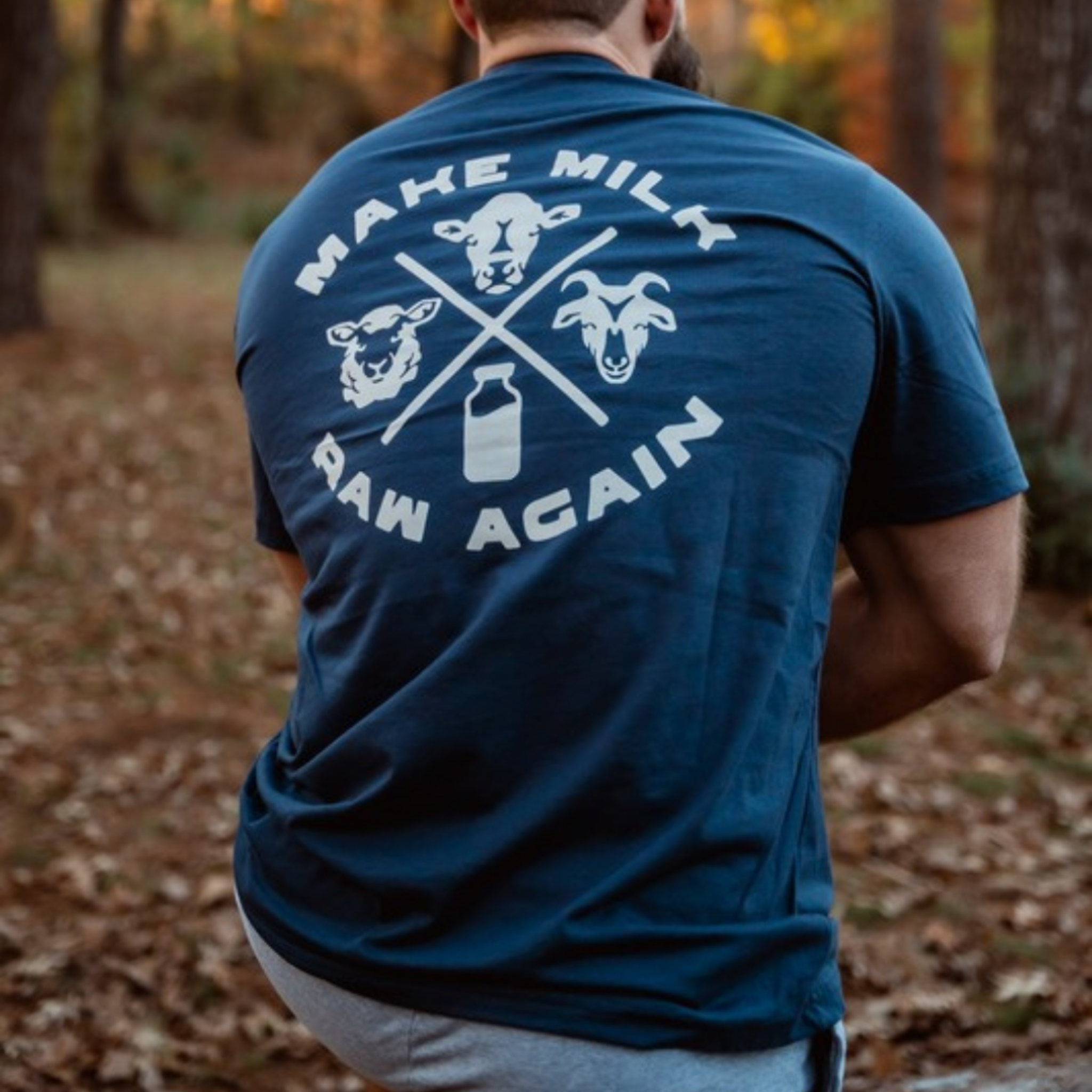 A person outdoors, seen from behind, wears the Farm Fresh Clothing MMRA Trinity Tee—an organic cotton shirt featuring MAKE MILK RAW AGAIN and graphics of a cow, sheep, goat, and milk bottle—with trees and fallen leaves in the background.