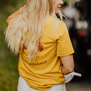 A woman with long blonde hair stands outdoors, facing away. She wears the yellow OG Bison Tee by Farm Fresh Clothing and light shorts, holding a white item in her left hand. The background features blurred greenery.