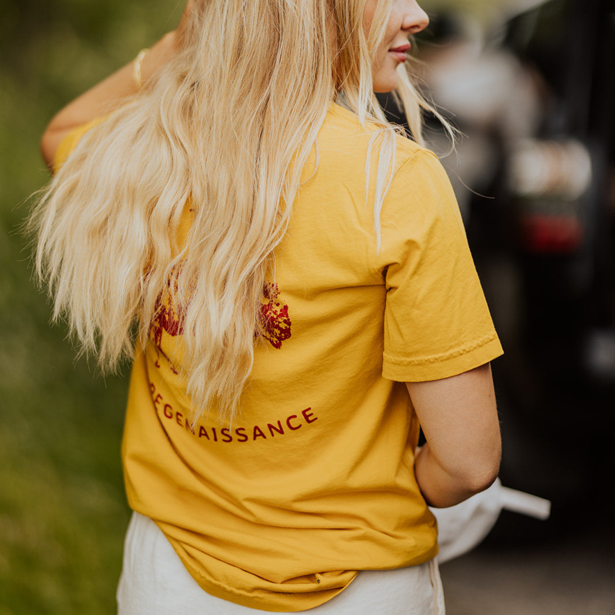 A woman with long blonde hair stands outdoors, facing away. She wears the yellow OG Bison Tee by Farm Fresh Clothing and light shorts, holding a white item in her left hand. The background features blurred greenery.