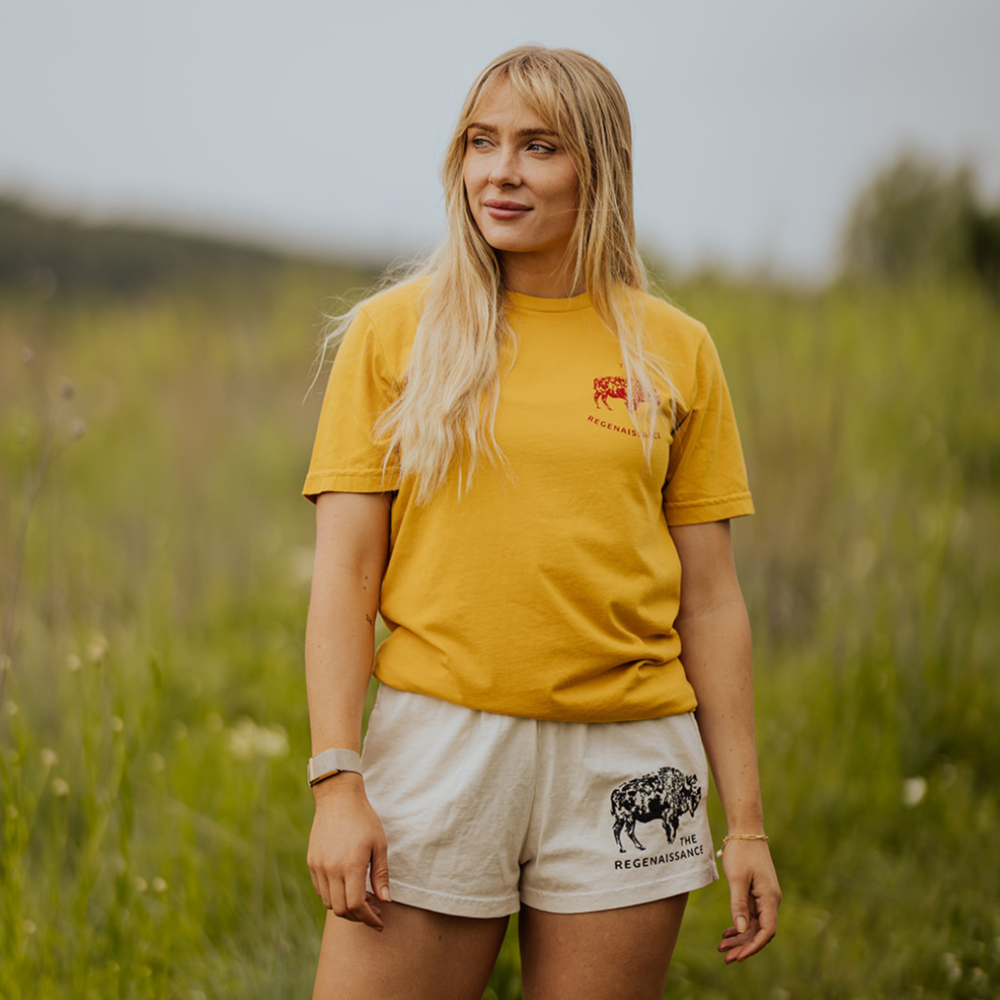 A woman with long blonde hair stands outdoors in a grassy field wearing the yellow OG Bison Tee by Farm Fresh Clothing and white shorts with a black "REGENASSANCE" graphic, gazing to the side with a neutral expression.