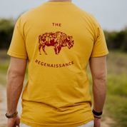 A person wearing the OG Bison Tee by Farm Fresh Clothing, featuring yellow organic cotton, a red bison graphic, and "THE REGENAISSANCE" on the back, stands outdoors on a path surrounded by greenery—a nod to regenerative agriculture.