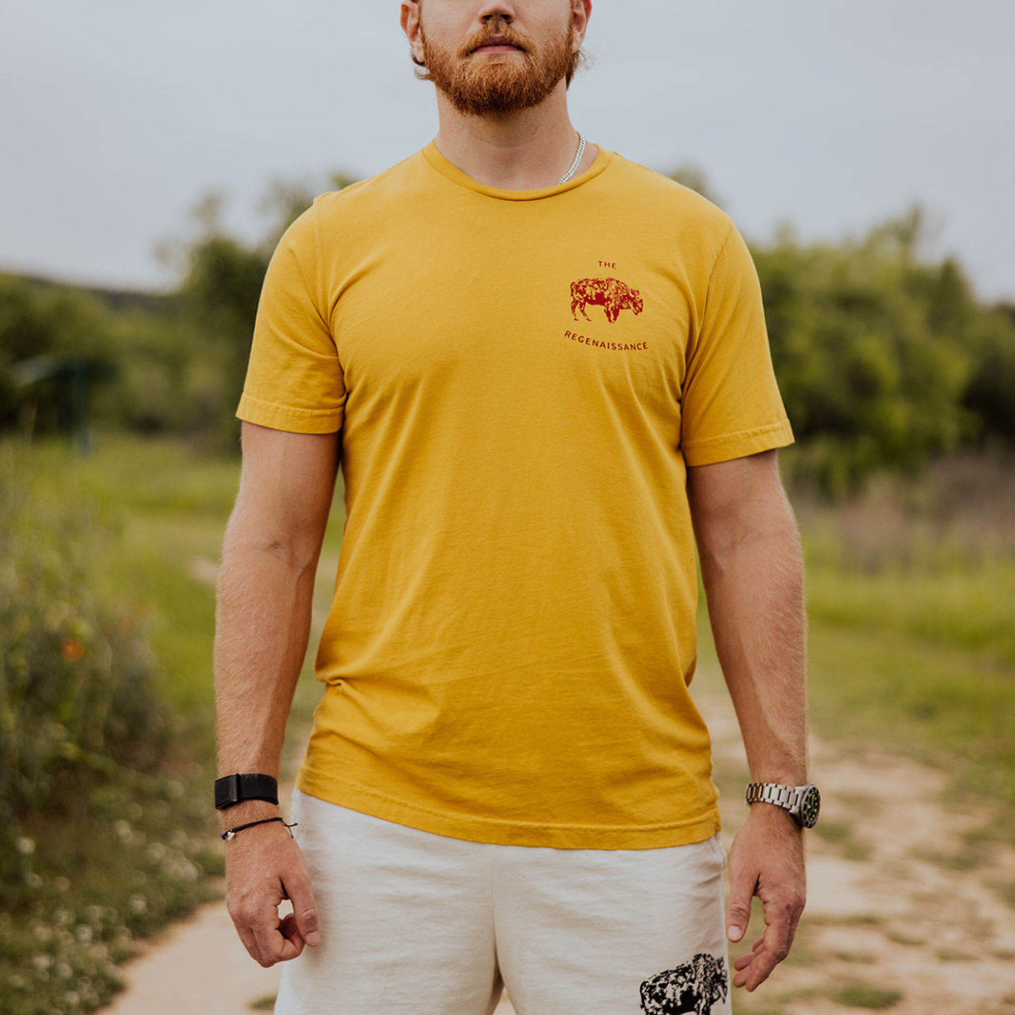 A bearded man stands outdoors in white shorts, wearing the Farm Fresh Clothing OG Bison Tee in mustard with a small red animal graphic and text on the chest, surrounded by greenery.