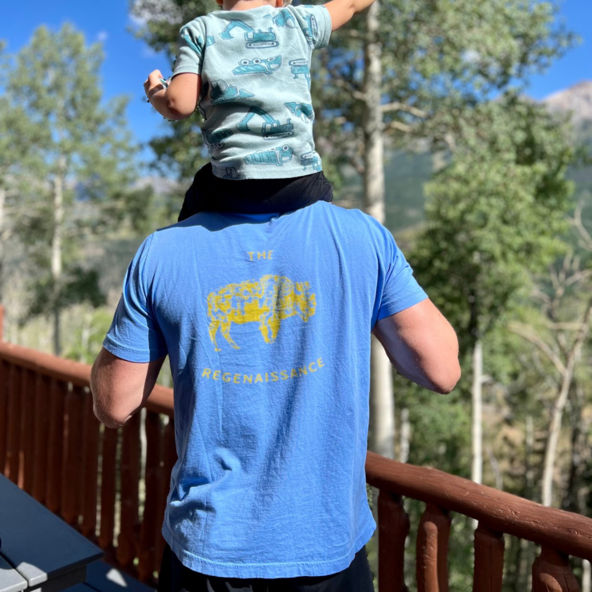 A man in a Farm Fresh Clothing OG Bison Tee stands on a wooden deck with a child on his shoulders, surrounded by trees and mountains.
