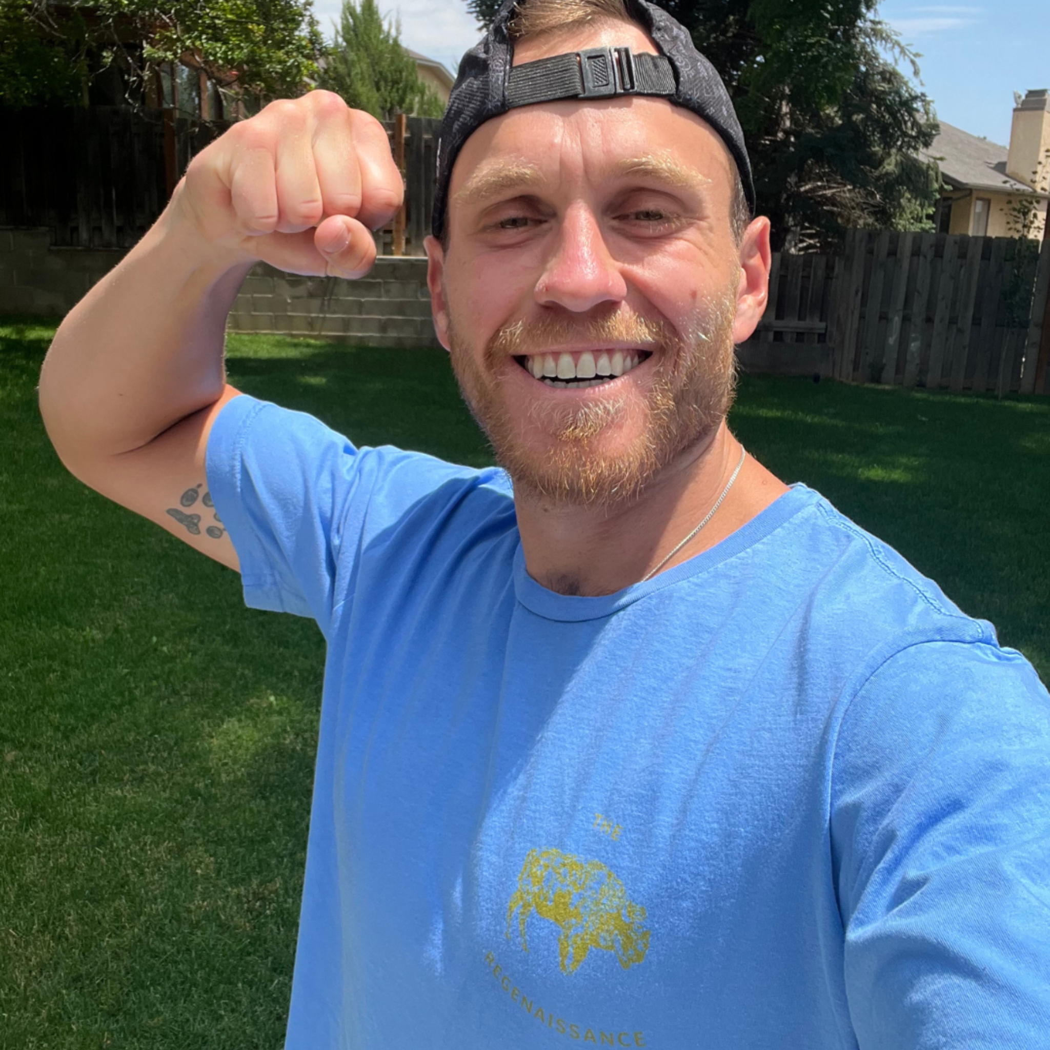 A man in the Farm Fresh Clothing OG Bison Tee—an organic cotton blue t-shirt—smiles and flexes his arm while wearing a black cap, standing in a sunny backyard with green grass and trees.