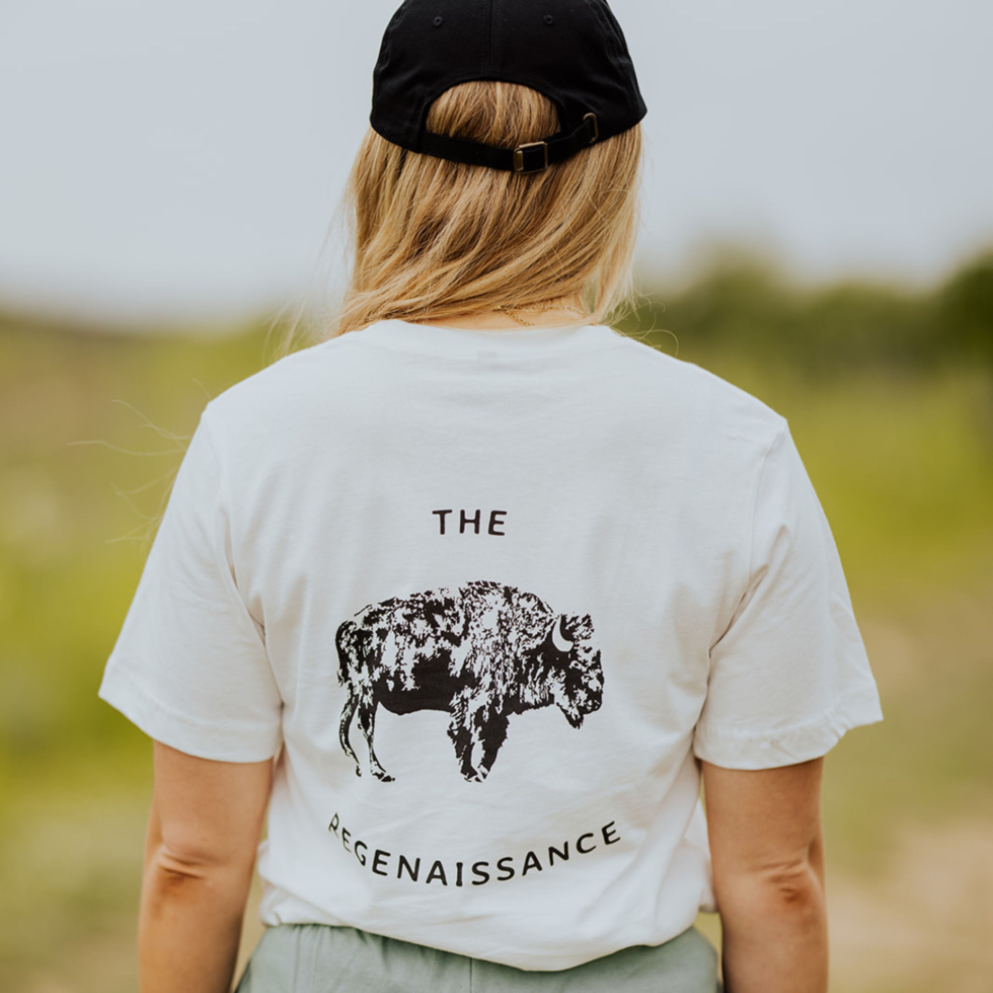 A person with long blonde hair and a black cap wears Farm Fresh Clothing’s OG Bison Tee, featuring “THE REGENAISSANCE,” while standing outdoors against a blurred green background.