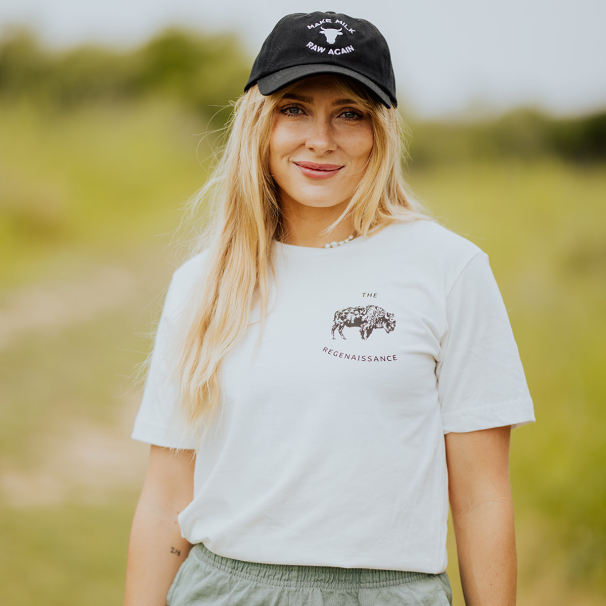 A woman with long blonde hair smiles outdoors on a grassy path, wearing a black cap, green pants, and the Farm Fresh Clothing OG Bison Tee—an organic cotton shirt featuring “The Regenaissance,” celebrating regenerative agriculture.