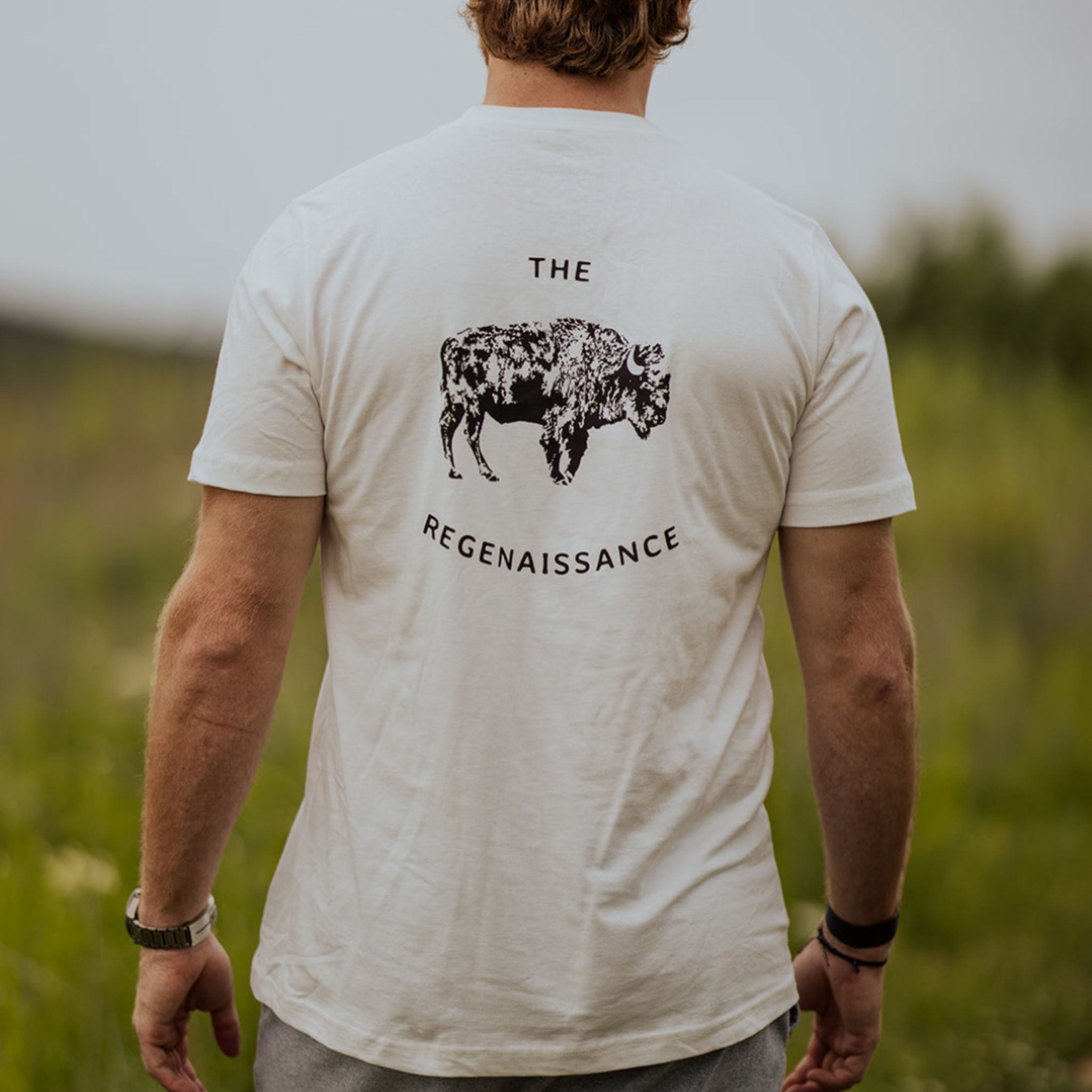 Seen from behind, a person stands outdoors on green grass wearing Farm Fresh Clothing’s OG Bison Tee, crafted from organic cotton and featuring a black bison illustration with THE REGENAISSANCE text on the back.