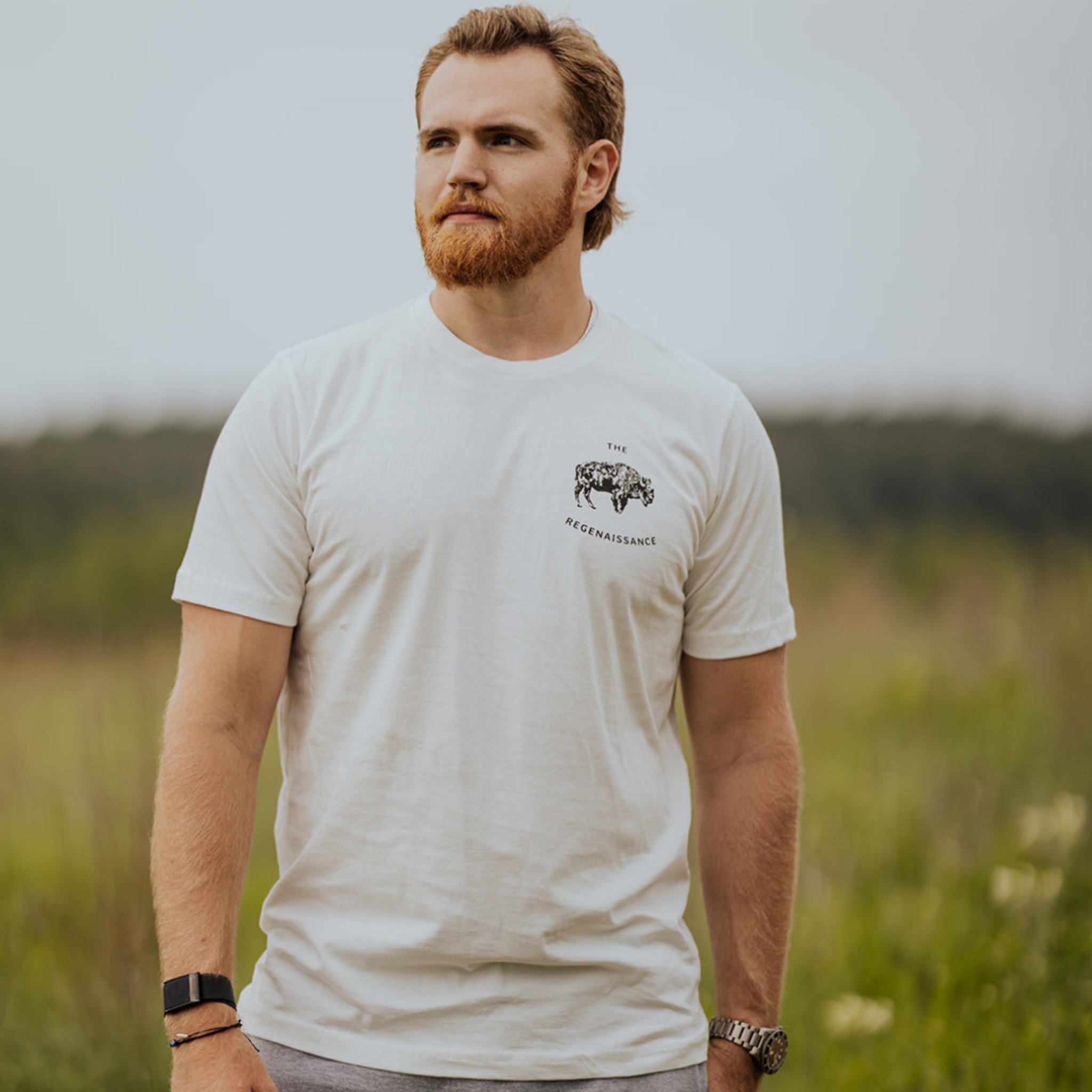A bearded man with short hair stands in a grassy field, wearing the Farm Fresh Clothing OG Bison Tee featuring a small graphic and text on the left chest. He accessorizes with a watch and bracelet; blurred greenery forms the background.
