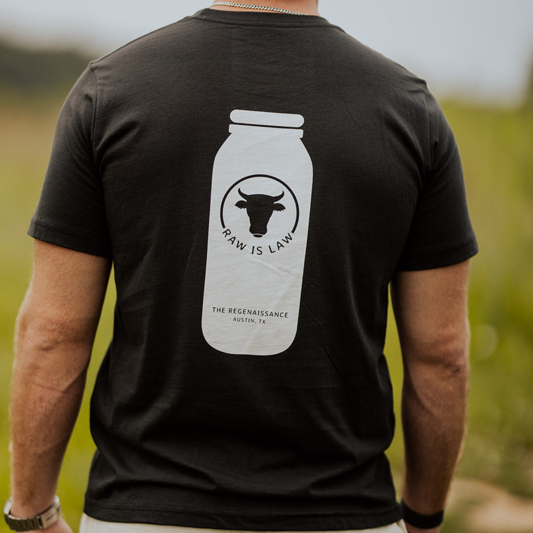 A person wears Farm Fresh Clothing’s Raw Is Law Tee, a black organic cotton shirt featuring a white milk bottle graphic, cow head, and “RAW IS LAW” text with “THE REGENAISSANCE AUSTIN, TX.” An outdoorsy setting highlights the theme of food freedom.