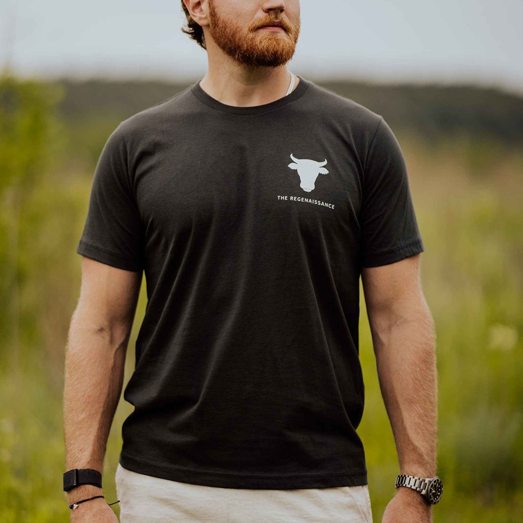 A bearded man stands outdoors in a Farm Fresh Clothing Raw Is Law Tee—an organic cotton black shirt featuring a white bull logo and "THE REGENAISSANCE" text on the left chest. He wears a watch, set against a blurred green backdrop.