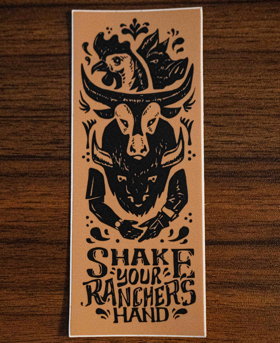 The Shake Your Ranchers Hand Sticker by Sticker Mule is a tan rectangular vinyl sticker featuring a cowboy hat with a chicken, a cow skull, and shaking hands, plus the message: SHAKE YOUR RANCHER’S HAND—know your farmer and choose real food.