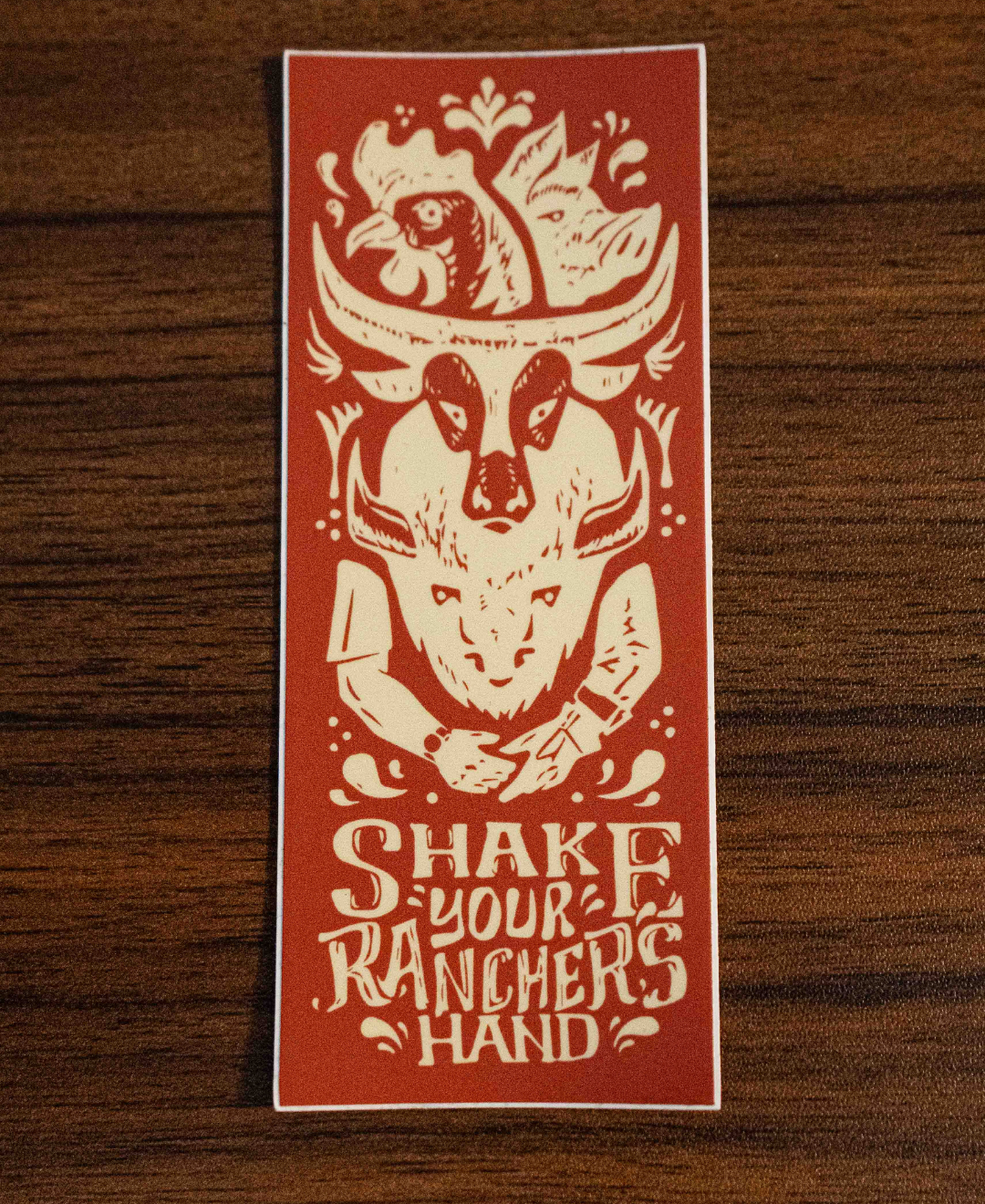 The Shake Your Ranchers Hand Sticker by Sticker Mule is a rectangular orange vinyl sticker featuring a chicken, steer skull, and hands about to shake, with SHAKE YOUR RANCHERS HAND text below in cream designs.