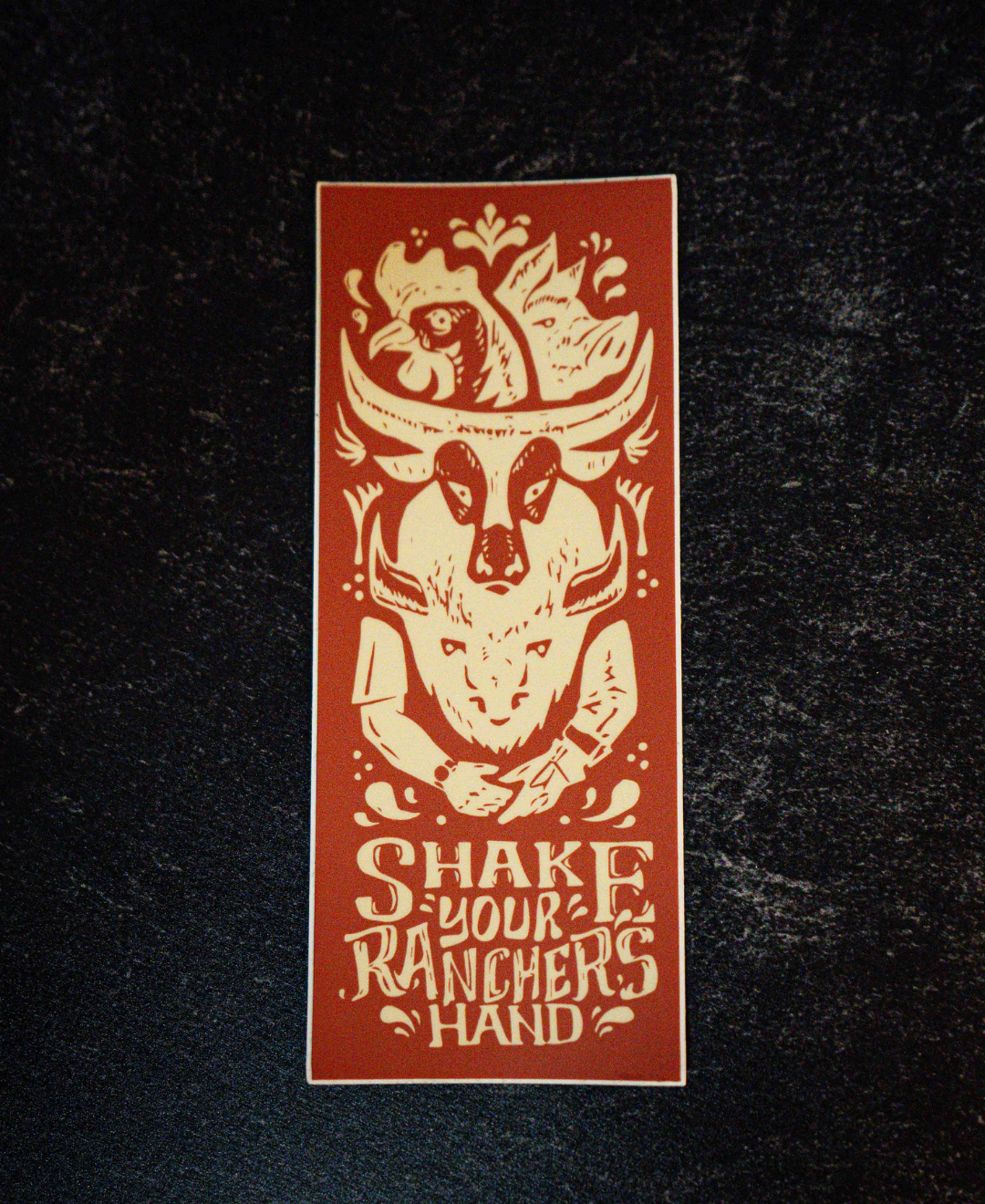 The Shake Your Ranchers Hand Sticker by Sticker Mule is a rectangular vinyl sticker with a cow skull, chicken, and handshake illustration, plus the phrase SHAKE YOUR RANCHERS HAND in brown and cream on a dark background.