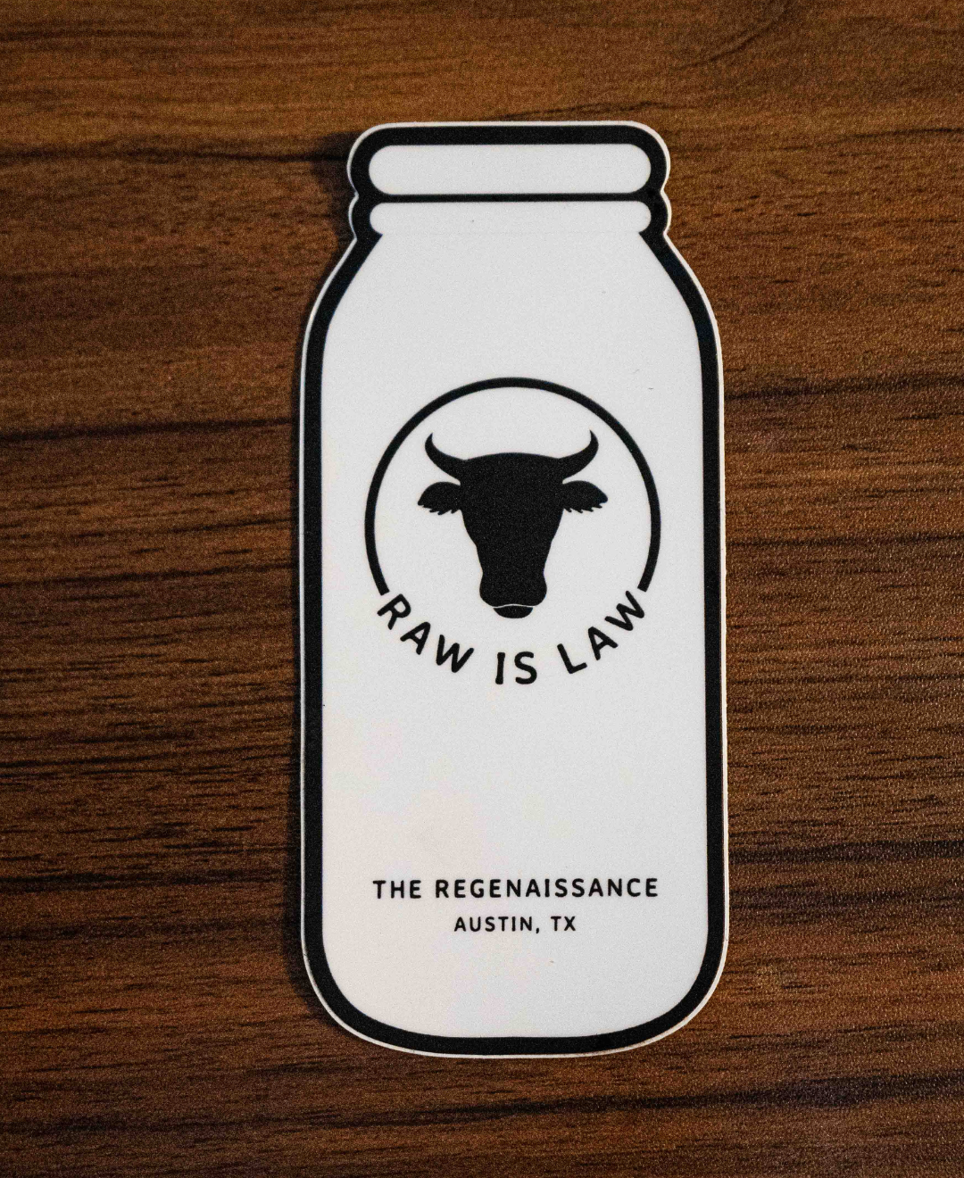 The Raw Is Law Sticker by Sticker Mule features a milk bottle with a cows head and RAW IS LAW text, plus THE REGENAISSANCE AUSTIN, TX below. Shown resting on a wooden surface, it highlights food rights.