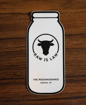 The Raw Is Law Sticker by Sticker Mule features a milk bottle with a cows head and RAW IS LAW text, plus THE REGENAISSANCE AUSTIN, TX below. Shown resting on a wooden surface, it highlights food rights.
