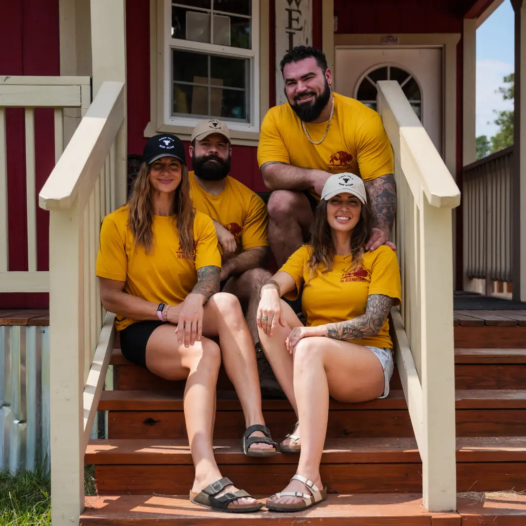 Group of two men and two women wearing mustard Regenerate Agriculture Tees with red bison logo, sitting together on farmhouse steps.