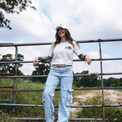 Woman smiling in eggshell Regenerate Agriculture Tee, front view, with navy bison and The Regenaissance logo, leaning on farm gate.