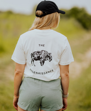 Wearing Los Angeles Apparel Women's Bison Shorts, a black cap, and a white "The Regenaissance" buffalo tee, a person stands on a grassy path outdoors, embodying regenerative agriculture and facing away from the camera.