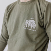 A person wears the Farm Fresh Clothing Bison Crewneck Sweatshirt in olive green, featuring an organic cotton build, a buffalo graphic, and THE REGENAISSANCE text; their face is partially out of frame.